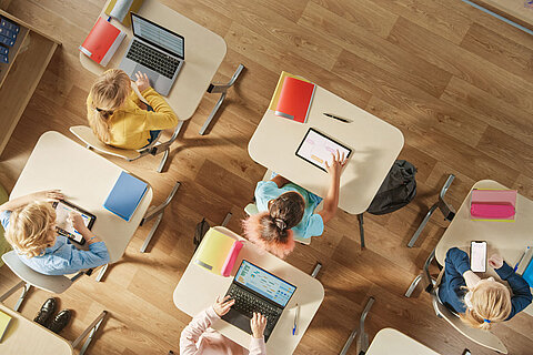 Top View Shot in Elementary School Computer Science Classroom: Children Sitting at their School Desk Using Personal Computers and Digital Tablets for Assignments.