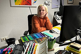 Eine Frau sitzt an einem Schreibtisch mit Buntstiften, Aquarellfarben, Kugelschreibern und Büromaterialien und blickt in die Kamera. Kunst- und Büromaterialien bedecken den Arbeitsbereich um sie herum.