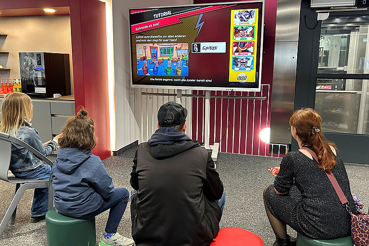 Vier Personen sitzen vor einem großen Bildschirm, auf dem eine bunte Spielanleitung in einem modernen Innenraum zu sehen ist.
