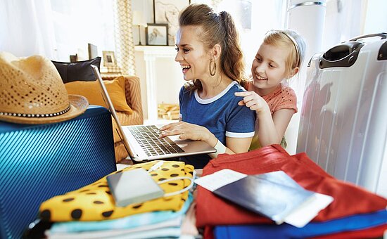 Eine Frau und ein Mädchen lächeln, während sie auf einen Laptop schauen, umgeben von Gepäck, Kleidung, einem Hut, einem Reisepass und Reisedokumenten, was darauf hindeutet, dass sie sich auf eine Reise vorbereiten.