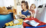 Eine Frau und ein Mädchen lächeln, während sie auf einen Laptop schauen, umgeben von Gepäck, Kleidung, einem Hut, einem Reisepass und Reisedokumenten, was darauf hindeutet, dass sie sich auf eine Reise vorbereiten.