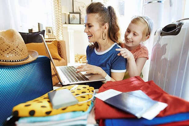 Eine Frau und ein Mädchen lächeln, während sie auf einen Laptop schauen, umgeben von Gepäck, Kleidung, einem Hut, einem Reisepass und Reisedokumenten, was darauf hindeutet, dass sie sich auf eine Reise vorbereiten.
