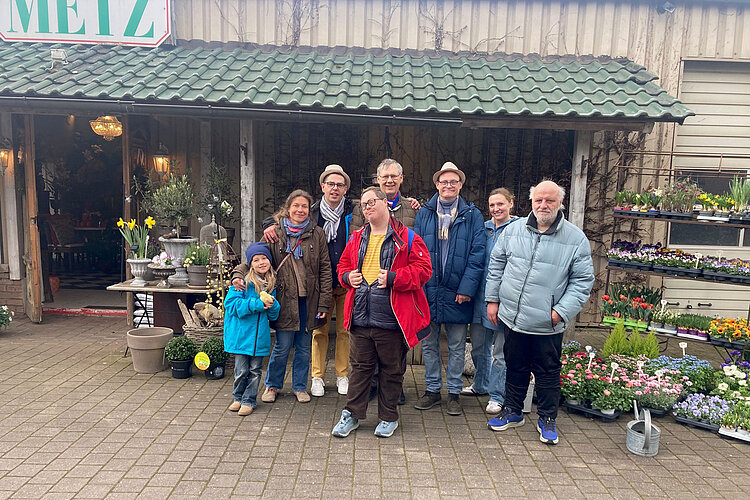 Eine Gruppe von acht Personen, darunter Erwachsene und Kinder, steht lächelnd vor einem Blumenladen, um den herum Topfpflanzen und bunte Blumen ausgestellt sind.