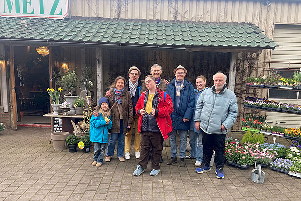 Eine Gruppe von acht Personen, darunter Erwachsene und Kinder, steht lächelnd vor einem Blumenladen, um den herum Topfpflanzen und bunte Blumen ausgestellt sind.
