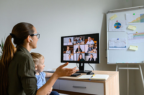 Back view of young mother in eyeglasses is talking online, she using lPC for video meeting with employees, cute baby boy sits on her laps. Busy mom works at home