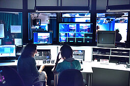 Zwei Personen sitzen in einem TV-Kontrollraum, umgeben von Bildschirmen, Monitoren und technischen Geräten, und beobachten Live-Übertragungen und verwalten eine Sendeproduktion.