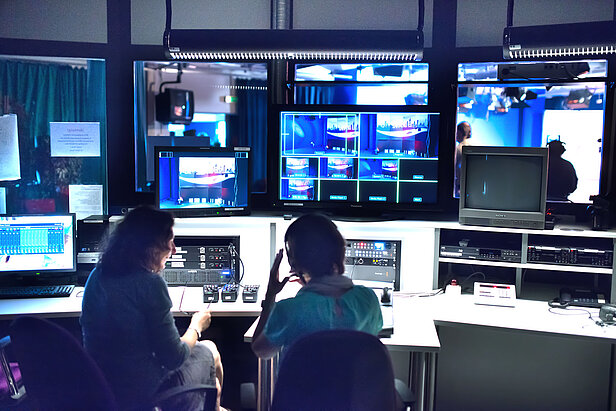 Zwei Personen sitzen in einem TV-Kontrollraum, umgeben von Bildschirmen, Monitoren und technischen Geräten, und beobachten Live-Übertragungen und verwalten eine Sendeproduktion.
