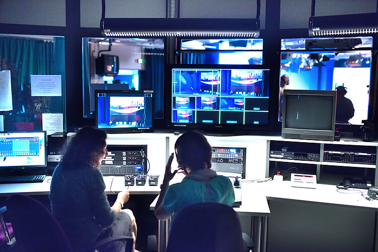 Zwei Personen sitzen in einem TV-Kontrollraum, umgeben von Bildschirmen, Monitoren und technischen Geräten, und beobachten Live-Übertragungen und verwalten eine Sendeproduktion.