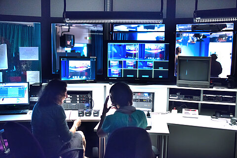 Zwei Personen sitzen in einem TV-Kontrollraum, umgeben von Bildschirmen, Monitoren und technischen Geräten, und beobachten Live-Übertragungen und verwalten eine Sendeproduktion.