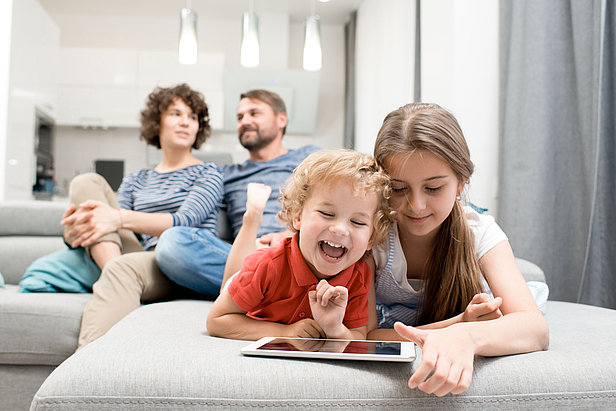 Portrait of brother and sister using digital tablet and laughing while lying on sofa in living room with parents watching TV in background