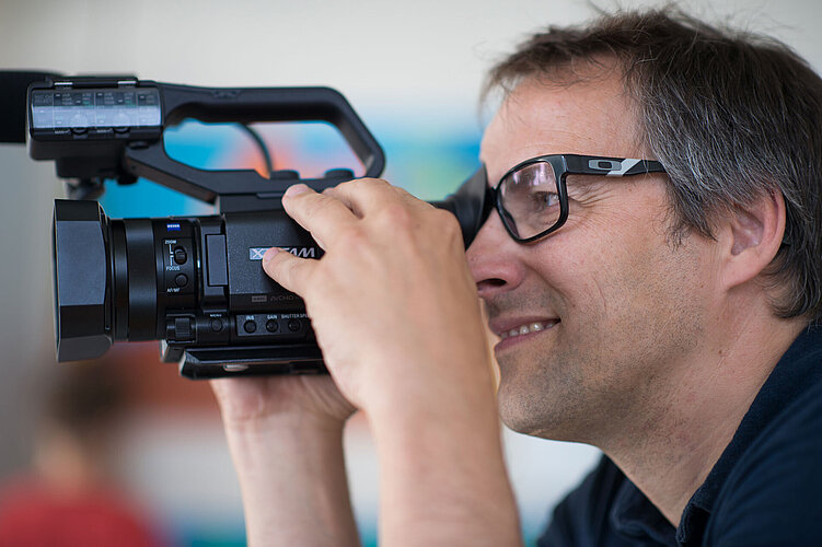 Eine Person mit Brille hält eine Videokamera in der Hand und blickt durch sie hindurch. Die Person ist konzentriert und lächelt leicht. Der unscharfe Hintergrund lässt auf eine Innenaufnahme schließen.