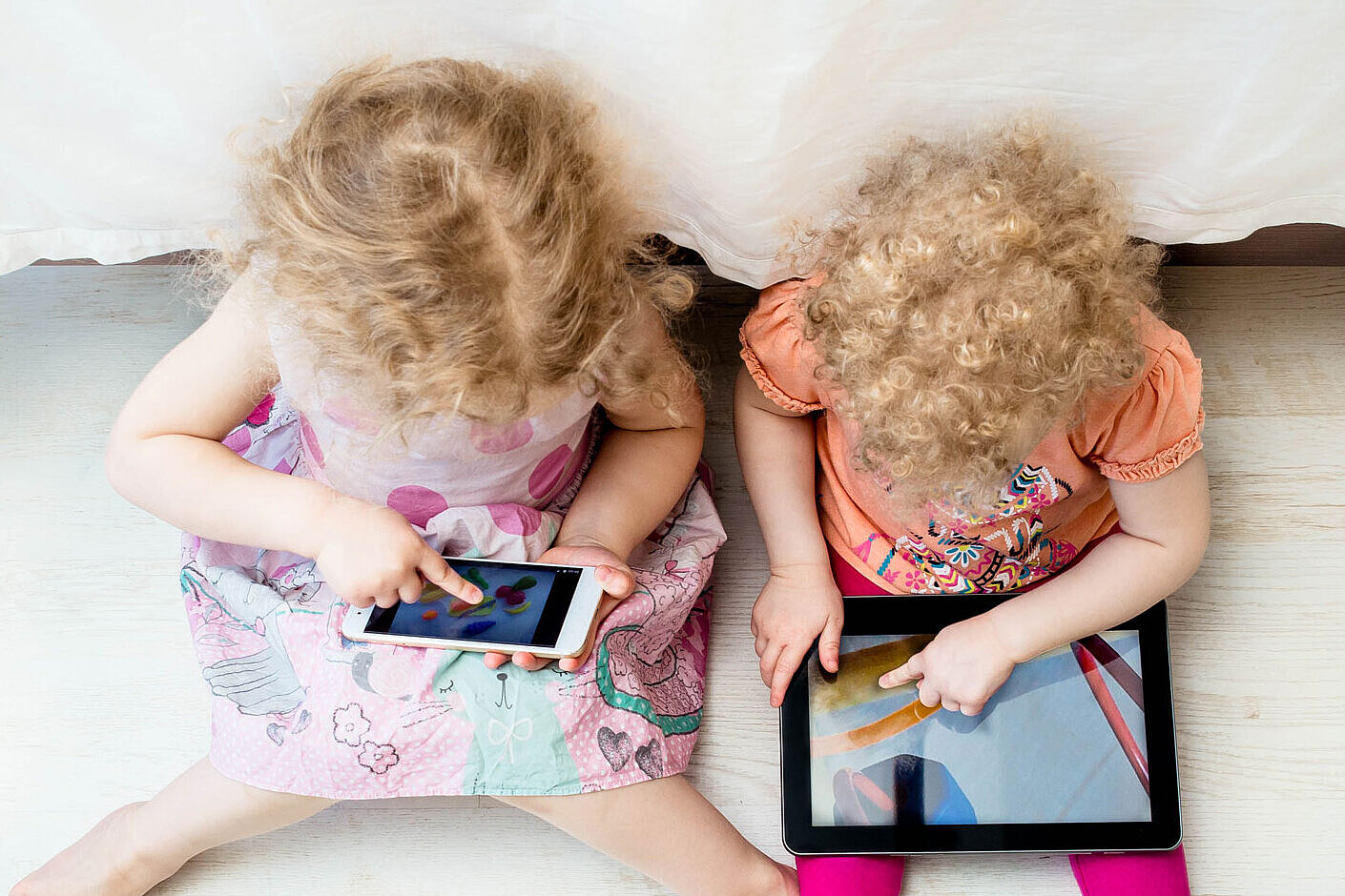 Zwei kleine Kinder mit lockigem Haar sitzen auf dem Boden und benutzen jeweils ein digitales Gerät. Ein Kind hält ein Smartphone in der Hand, während das andere ein Tablet benutzt. Beide sind auf ihre Bildschirme konzentriert und sitzen nebeneinander neben einem Bett.