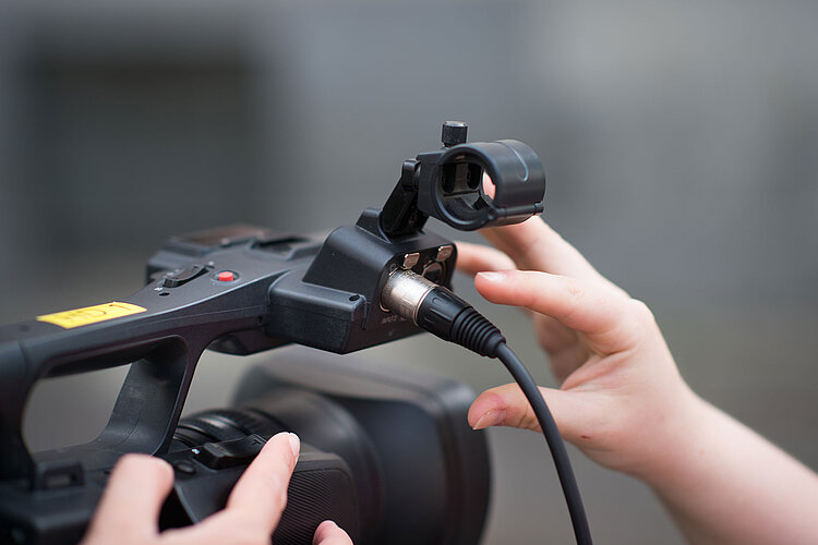 Nahaufnahme der Hände einer Person, die ein Mikrofonkabel an eine professionelle Videokamera anschließt, wobei der Fokus auf dem Anschluss und den Bedienelementen der Kamera liegt. Der Hintergrund ist unscharf.