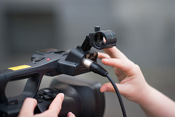 Nahaufnahme der Hände einer Person, die ein Mikrofonkabel an eine professionelle Videokamera anschließt, wobei der Fokus auf dem Anschluss und den Bedienelementen der Kamera liegt. Der Hintergrund ist unscharf.