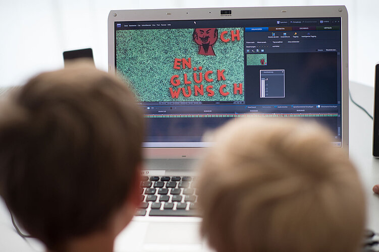 Zwei Kinder betrachten einen Laptop-Bildschirm, auf dem eine Videobearbeitungssoftware läuft. Das Video zeigt das Wort „GLÜCKWUNSCH“ und einen Teil eines roten Designs auf grünem Hintergrund. Der Fokus liegt auf dem Bildschirm, die Kinder sind im Vordergrund unscharf dargestellt.