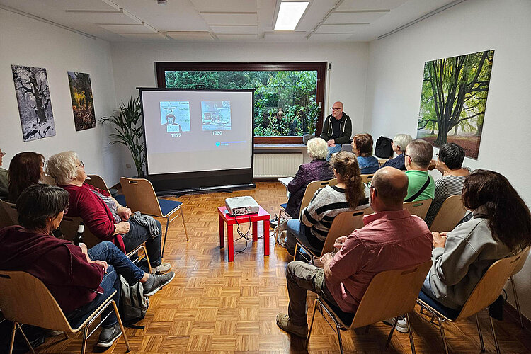 Eine Gruppe von Menschen sitzt in einem kleinen Raum und verfolgt eine Präsentation, die auf eine Leinwand projiziert wird.