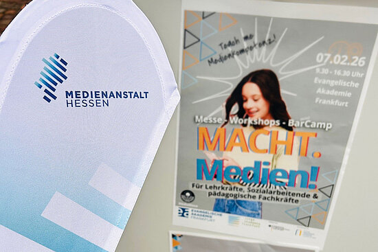 Beachflag Medienanstalt Hessen vor einem Plakat für die Veranstaltung "MACHT. Medien!" in der Evangelischen Akademie Frankfurt, das eine lächelnde Frau mit Schrift auf dem Plakat zeigt.