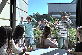 Eine Gruppe Kinder sitzt drinnen mit Getränken an einem Tisch, lächelt und beobachtet drei Jungen, die an einem sonnigen Tag draußen winkend vor dem Fenster stehen.