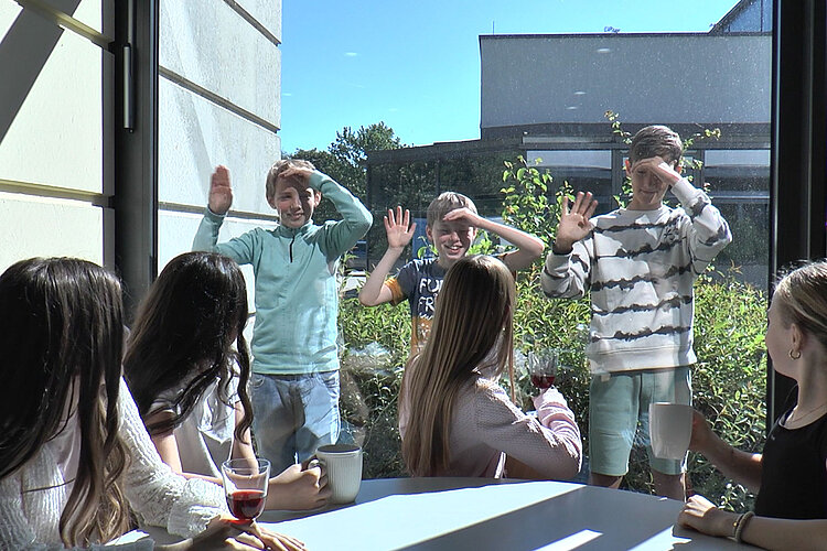 Eine Gruppe Kinder sitzt drinnen mit Getränken an einem Tisch, lächelt und beobachtet drei Jungen, die an einem sonnigen Tag draußen winkend vor dem Fenster stehen.