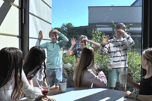 Eine Gruppe Kinder sitzt drinnen mit Getränken an einem Tisch, lächelt und beobachtet drei Jungen, die an einem sonnigen Tag draußen winkend vor dem Fenster stehen.