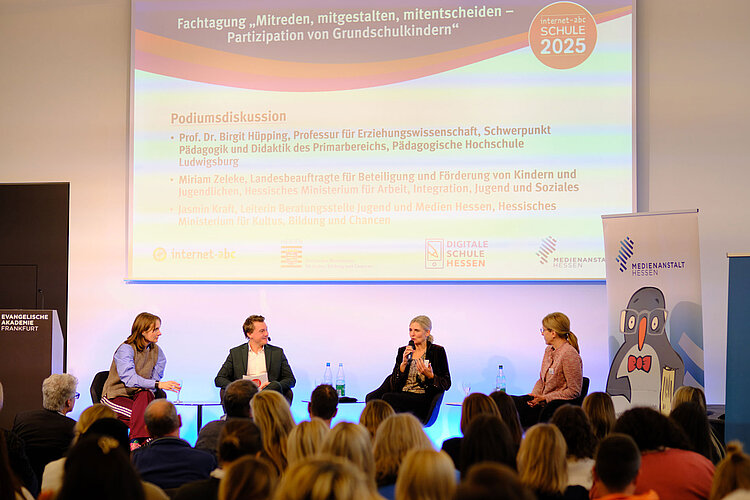 Vier Podiumsteilnehmer und ein Moderator sitzen auf der Bühne und diskutieren über die Beteiligung von Grundschülern. Auf einem Bildschirm hinter ihnen werden das Thema der Veranstaltung und die Namen der Diskussionsteilnehmer angezeigt. Das Publikum schaut zu, und an den Seiten sind Banner mit Sponsorenlogos zu sehen.