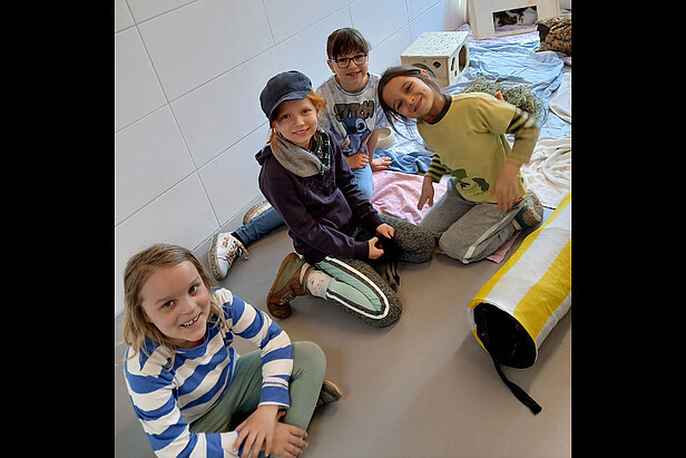 Vier lächelnde Kinder sitzen in einem hellen Raum mit weißen Wänden auf dem Boden, umgeben von Decken, einem Tunnelspielzeug und einem Haustierkorb. 
