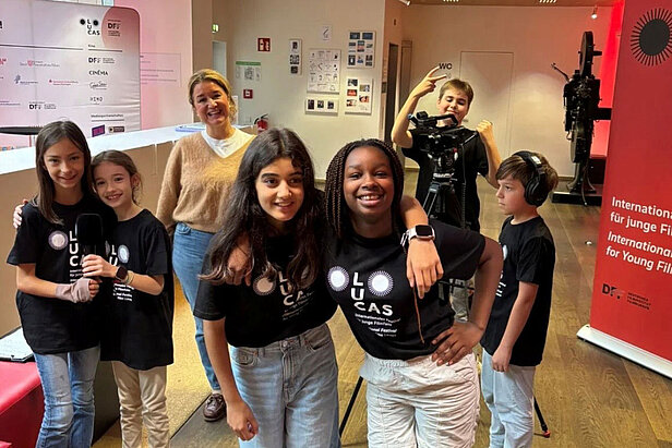 Eine Gruppe lächelnder Kinder und ein Erwachsener posieren in einem Haus. Vier Kinder tragen passende schwarze "LUCAS"-Shirts, und ein Kind bedient eine Videokamera. Im Hintergrund sind Plakate und ein großer roter Bildschirm zu sehen.
