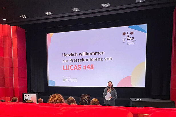 Eine Person steht auf der Bühne und hält ein Mikrofon vor einer Leinwand mit der Aufschrift "Herzlich willkommen zur Pressekonferenz von LUCAS #48" in einem rot-schwarzen Kinosaal, der mit sitzenden Teilnehmern gefüllt ist.