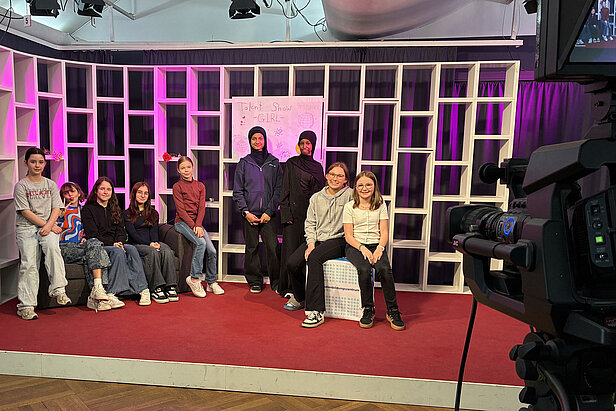 Eine Gruppe von Kindern sitzt und steht auf einer Bühne mit einem roten Teppich und weißen Regalen im Hintergrund. Hinter ihnen hängt ein Schild mit der Aufschrift "Talent Show GIRLS". Im Vordergrund rechts ist eine Kamera zu sehen.