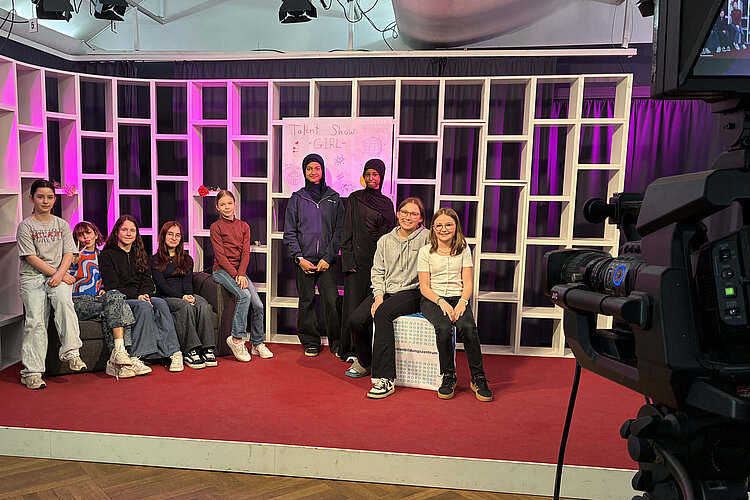 Eine Gruppe von Kindern sitzt und steht auf einer Bühne mit einem roten Teppich und weißen Regalen im Hintergrund. Hinter ihnen hängt ein Schild mit der Aufschrift "Talent Show GIRLS". Im Vordergrund rechts ist eine Kamera zu sehen.