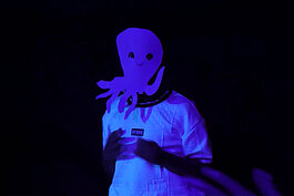 Eine Person in weißer Kleidung und mit einer großen, leuchtend lila Krakenmaske steht vor einem dunklen Hintergrund, der von Schwarzlicht beleuchtet wird.
