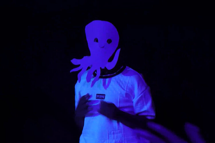Eine Person in weißer Kleidung und mit einer großen, leuchtend lila Krakenmaske steht vor einem dunklen Hintergrund, der von Schwarzlicht beleuchtet wird.