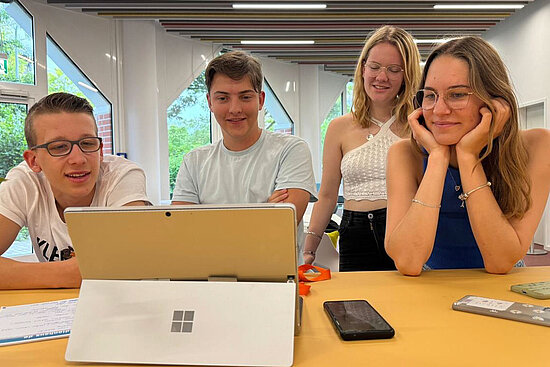 Vier junge Leute sitzen und stehen um einen Tisch herum, lächeln und schauen auf einen Laptop. Auf dem Tisch liegen Papiere, ein Smartphone und Stifte.