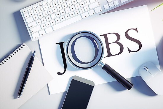 Ein Schreibtisch mit einer Tastatur, einer Maus, einem Smartphone, einer Tasse Kaffee und einer Pflanze. Eine Lupe liegt über einem Blatt Papier, auf dem das Wort „JOBS“ aufgedruckt ist.