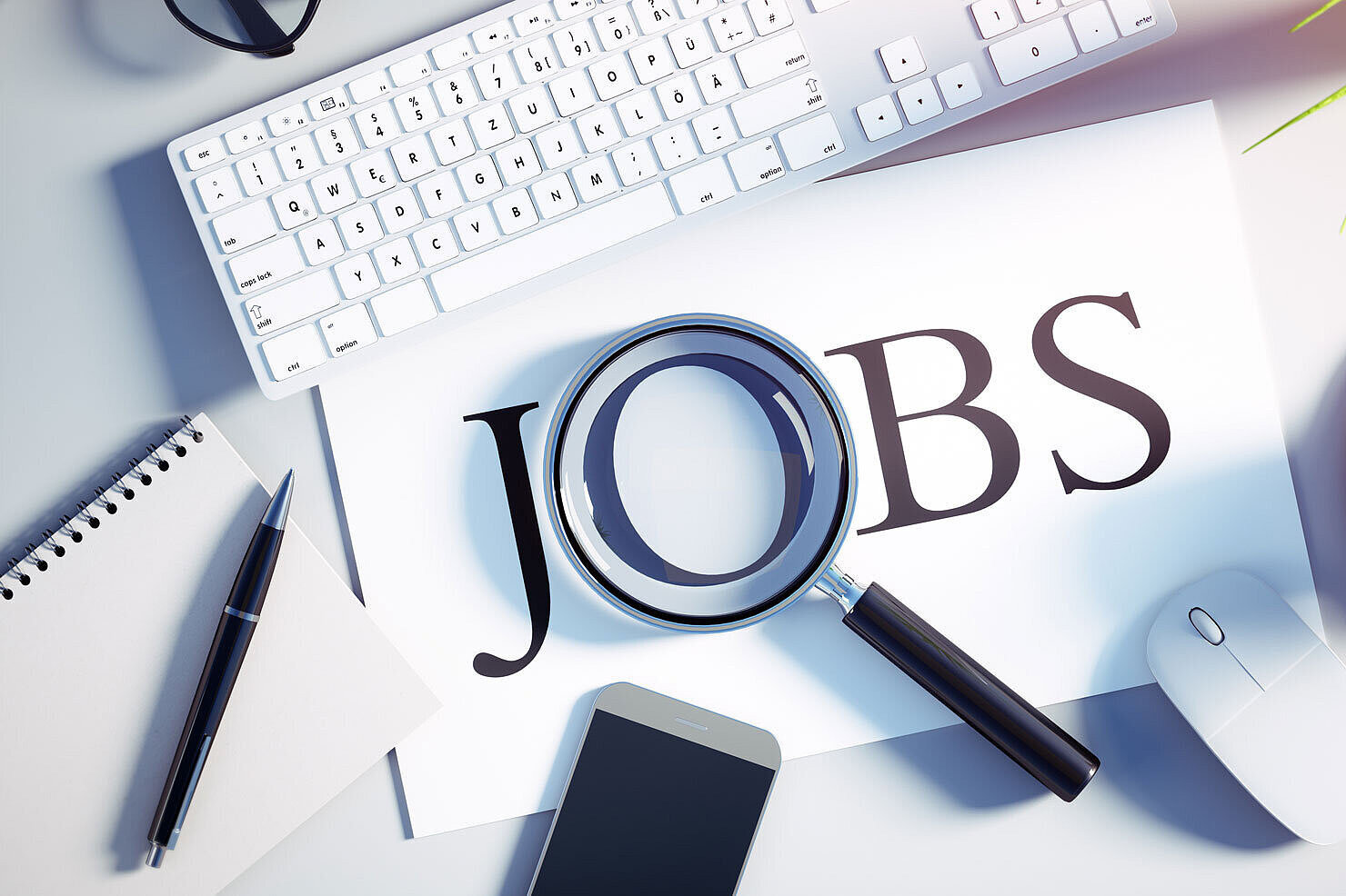 Ein Schreibtisch mit einer Tastatur, einer Maus, einem Smartphone, einer Tasse Kaffee und einer Pflanze. Eine Lupe liegt über einem Blatt Papier, auf dem das Wort „JOBS“ aufgedruckt ist.