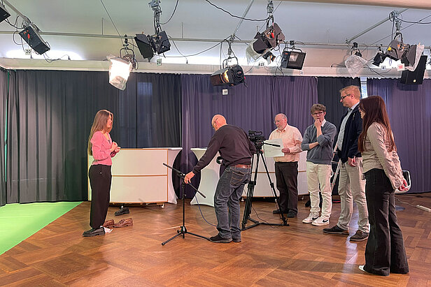 Eine Frau steht vor einer Kamera und einem Mikrofon in einem hell erleuchteten Studio, während vier Männer und eine Frau die Ausrüstung einstellen und die Einrichtung beobachten. Im Hintergrund sind ein Greenscreen und Vorhänge zu sehen.