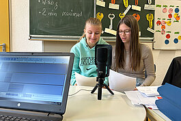 Zwei Mädchen im Teenageralter sitzen zusammen an einem Tisch mit Papieren und sprechen in ein Mikrofon. Im Vordergrund ist ein Laptop mit Audiosoftware aufgeklappt. Hinter ihnen sind Kreidetafeln mit Schrift und bunten Tasten zu sehen.