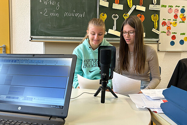 Zwei Mädchen im Teenageralter sitzen zusammen an einem Tisch mit Papieren und sprechen in ein Mikrofon. Im Vordergrund ist ein Laptop mit Audiosoftware aufgeklappt. Hinter ihnen sind Kreidetafeln mit Schrift und bunten Tasten zu sehen.