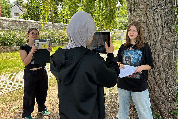 Drei Mädchen stehen im Freien in der Nähe eines Baumes; eine interviewt eine andere, die Papiere in der Hand hält, während eine dritte sie mit einem Tablet filmt. Im Hintergrund sind eine Steinmauer und Grünflächen zu sehen.