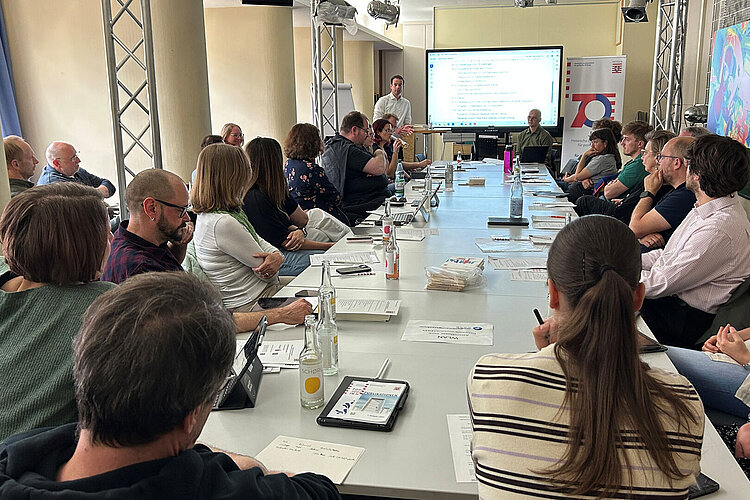 Mehrere Personen sitzen an einem Konferenztisch und verfolgen eine Präsentation auf einem Bildschirm