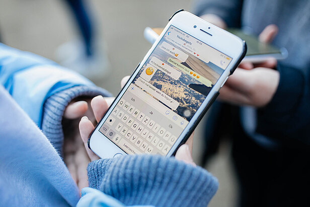 Eine Person hält ein Smartphone, auf dem eine Messaging-App mit einer sichtbaren Konversation angezeigt wird. Sie trägt eine blau-graue Jacke. Im Hintergrund hält eine andere Person ein Telefon.