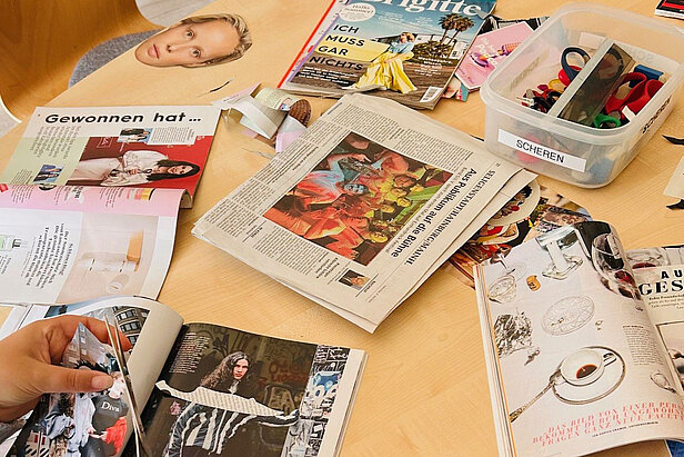 Eine Tischplatte mit aufgeschlagenen Zeitschriften, einer Zeitung, Bastelmaterial, Scheren und ausgeschnittenen Bildern, wobei jemand eine Zeitschriftenseite in der Hand hält.