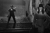 Eine Frau in Vintage-Kleidung und mit Hut steigt mit einem Koffer in der Hand eine dunkle Treppe hinauf. Im Hintergrund steht eine Silhouette mit Hut oben auf der Treppe und hält einen Geigenkasten in der Hand. Dies schafft eine dramatische Szene im Stil eines Film Noir.