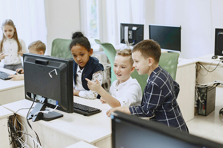 Drei Kinder sitzen in einem Klassenzimmer um einen Desktop-Computer herum, schauen auf den Bildschirm und diskutieren etwas, während andere Schüler an den benachbarten Tischen arbeiten.
