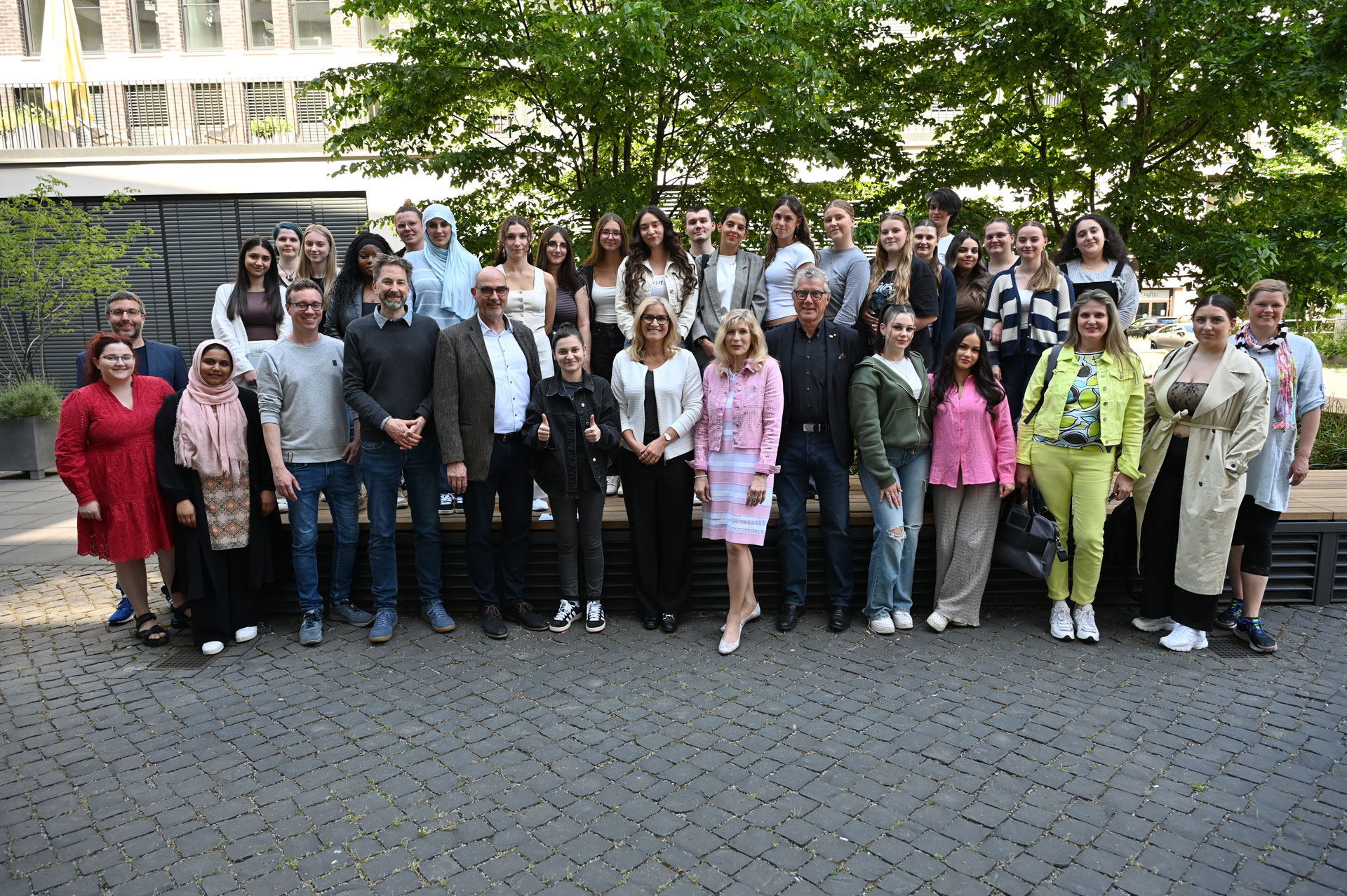 Eine große Gruppe von Menschen, darunter Männer und Frauen unterschiedlichen Alters, steht draußen auf einem Kopfsteinpflasterweg zusammen und posiert und lächelt für ein Gruppenfoto vor grünen Bäumen und einem modernen Gebäude.