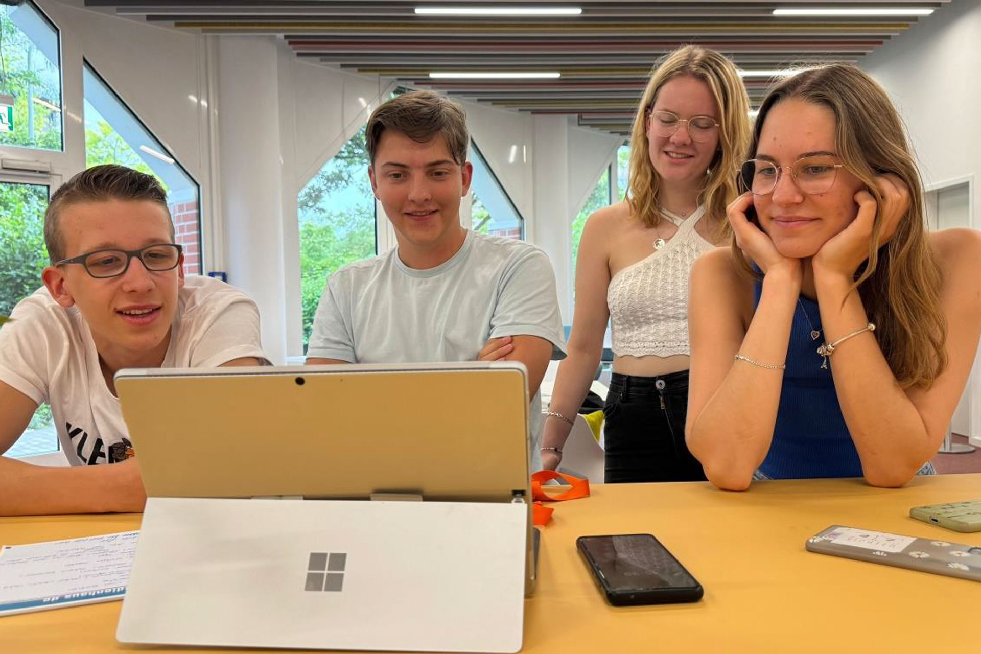 Vier junge Leute sitzen und stehen um einen Tisch herum, lächeln und schauen auf einen Laptop. Auf dem Tisch liegen Papiere, ein Smartphone und Stifte.