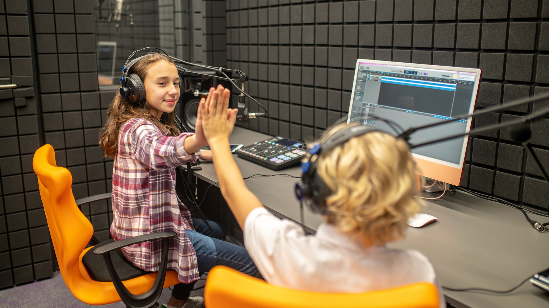 Zwei Kinder mit Kopfhörern sitzen in einem Aufnahmestudio an einem Tisch mit Mikrofonen, einem Mischpult und einem Computermonitor, auf dem eine Audiobearbeitungssoftware angezeigt wird, und klatschen sich gegenseitig ab.