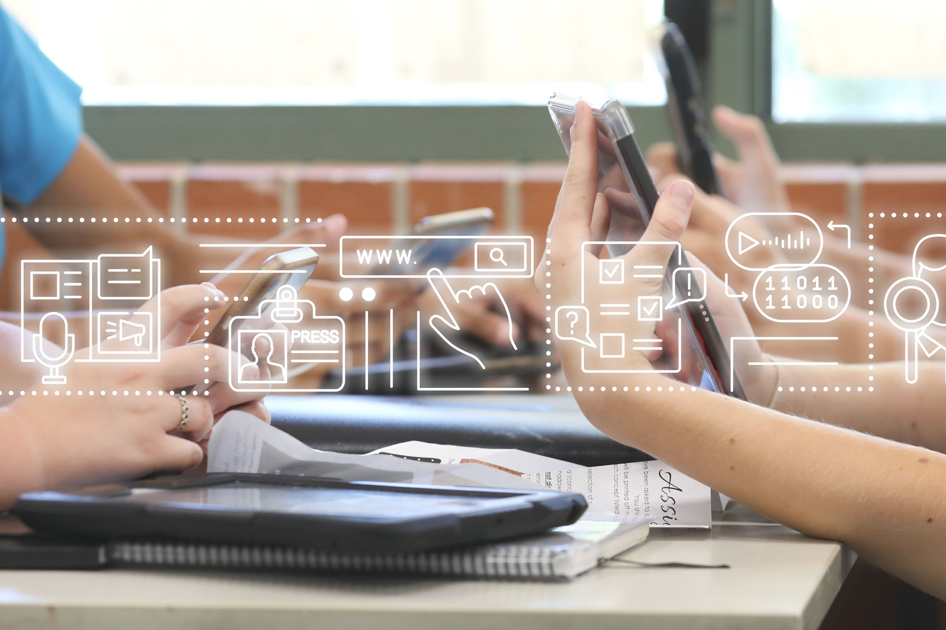 A table of high school students all holding multiple digital devices such as phones and tablets during a class lesson.